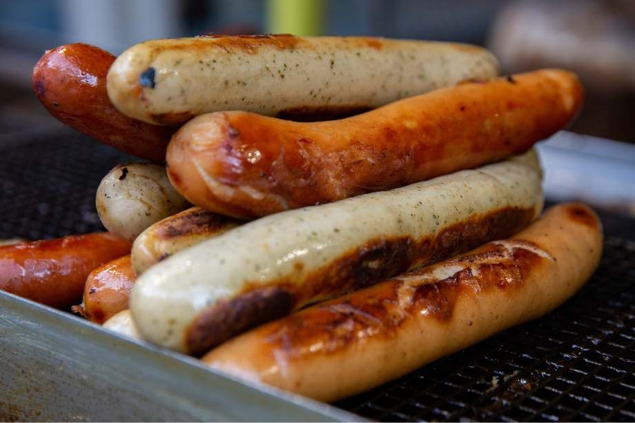 Pile of cooked hot dogs on a large grill with buns in the background.