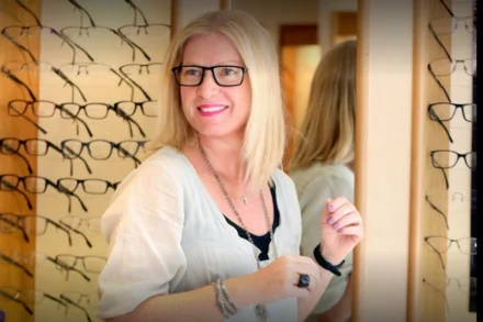 Inside of shop with racks of glasses on the wall with blonde female in the foreground wearing glasses and smiling.